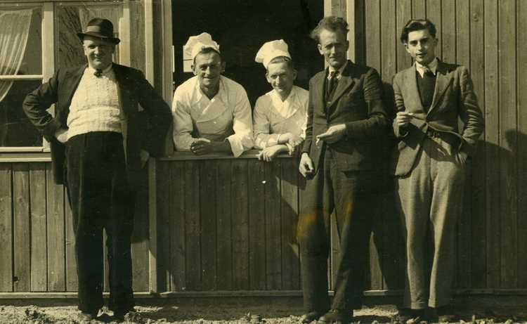 Personeel van kamp Vollenhove poseert op een mooie dag tegen de buitenmuur van een barak. V.l.n.r. kampbeheerder Appels, kok Jan van Urk, een tweede kok en twee kantoorklerken, ca. 1947 (privécollectie familie Appels)