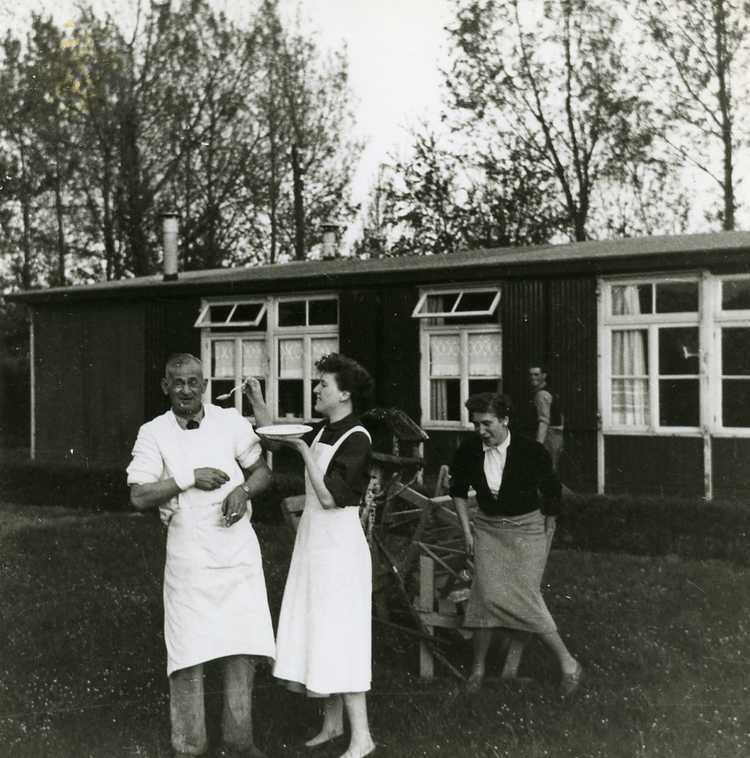 Martha en Lofa Appels dollen met kok Frans de Graaf in kamp Schokland. Op de achtergrond het huis van de familie Appels, ca. 1958 (privécollectie familie Appels)