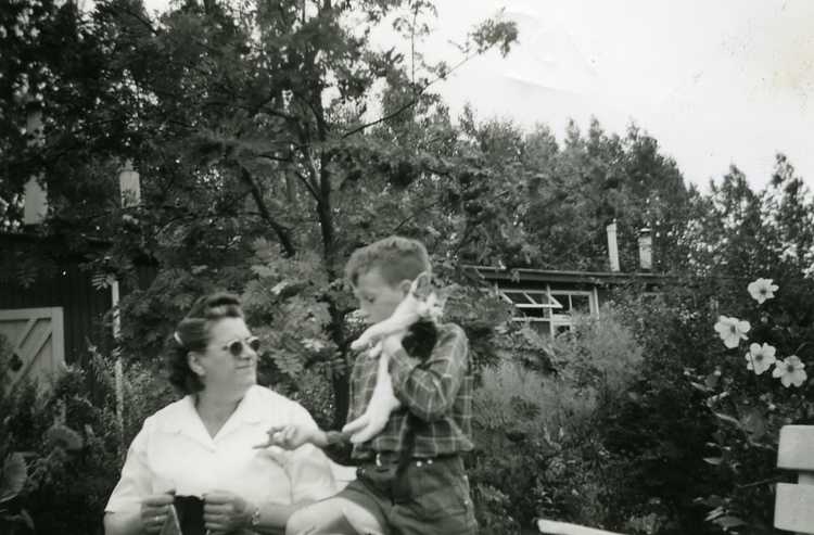 Moeder Appels, altijd met een breiwerk, en broertje Peter in de weelderige tuin van kamp Schokland. Op de achtergrond een arbeidersbarak, ca. 1955 (privécollectie familie Appels)