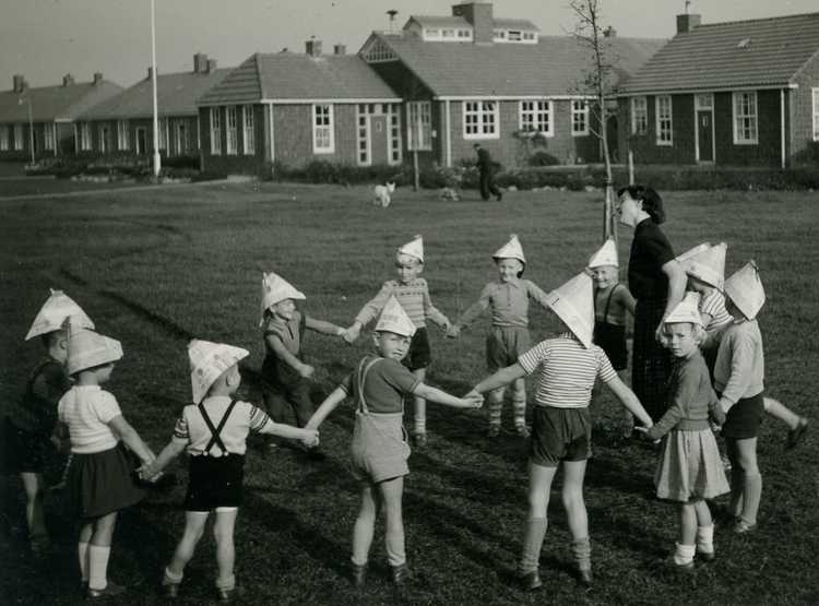 De eerste daarvoor gebouwde school had drie lokalen. Het kleuterlokaal was het meest noordelijke. Hier dansen de kleuters met juf Blomkwist op het grasveld van het stenen kamp, 1956